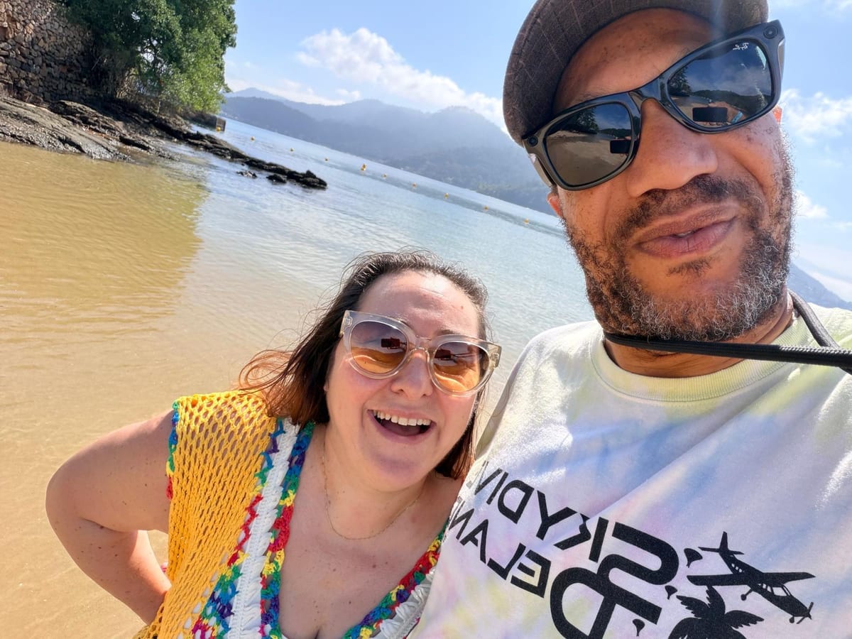 Delphine et Pedro, souriants côte à côte sur une plage, la mer calme et des collines boisées derrière eux